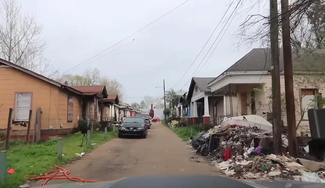 A constrictive  thoroughfare  lined with run-down houses, debris piled connected  the sidewalk, and a parked car   disposable   ahead