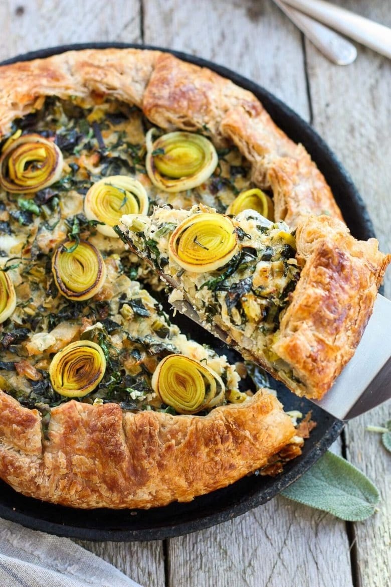 Savory vegetable and cheese galette with a slice being served, featuring artfully arranged leeks and flaky, golden crust