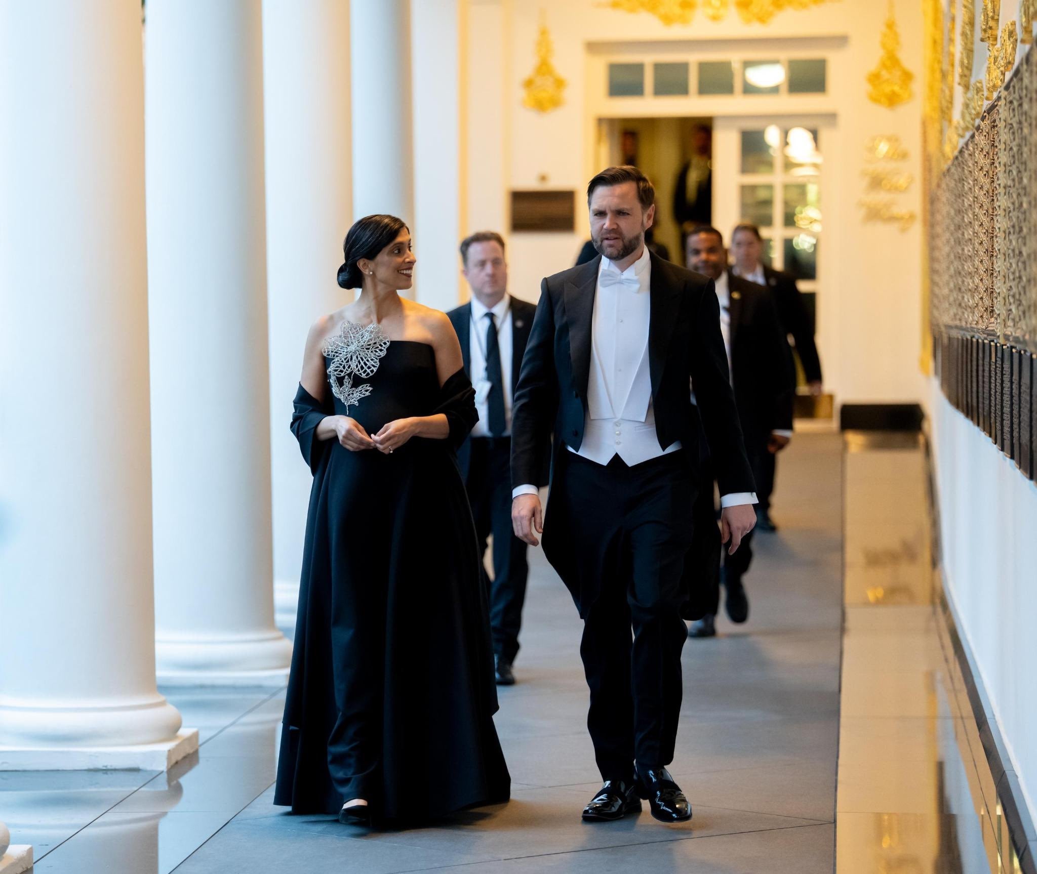 A man and a woman in formal attire walk down a hallway with decorative columns. The woman wears a black gown with an ornate shoulder design