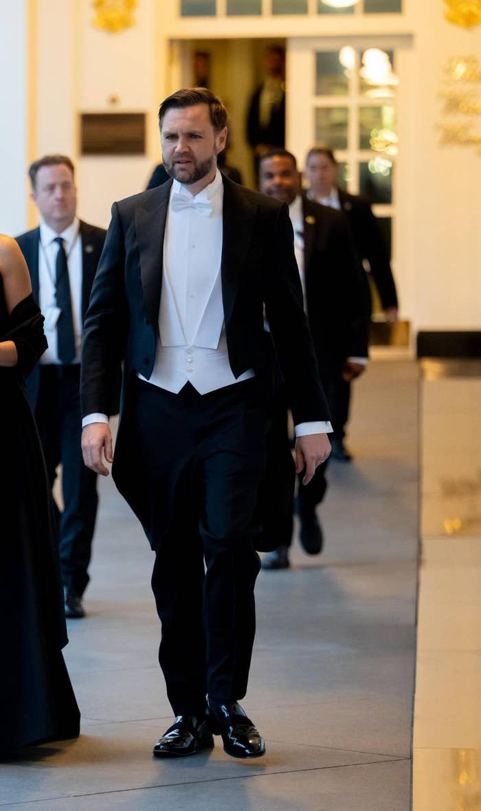 A man and a woman in formal attire walk down a hallway with decorative columns. The woman wears a black gown with an ornate shoulder design
