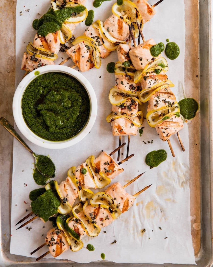 Grilled salmon skewers with lemon slices and herbs served with a bowl of green sauce on parchment paper