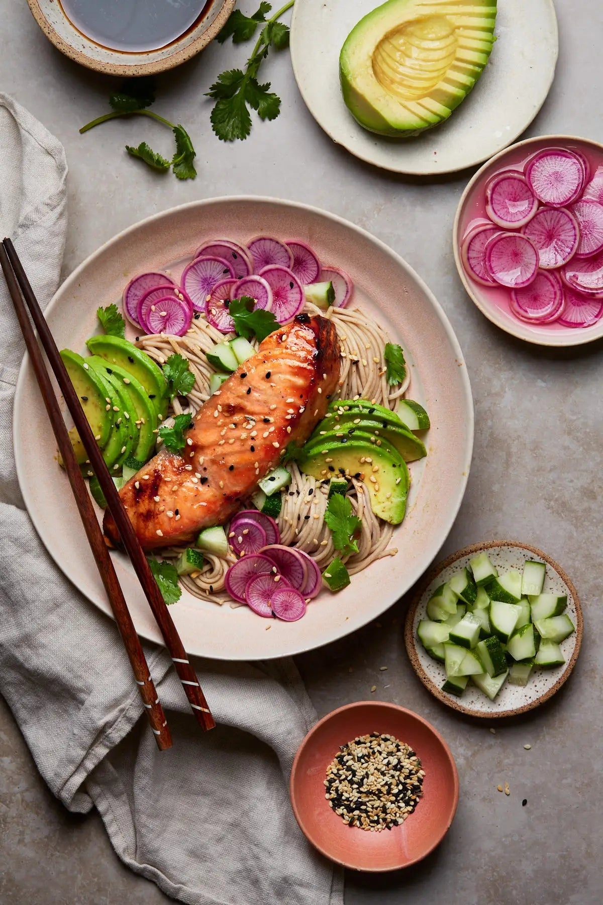 A bowl of soba noodles topped with glazed salmon, avocado slices, radishes, and cilantro, with small bowls of cucumber, radishes, and seeds nearby