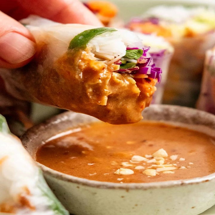 A hand holding a fresh spring roll above a bowl of peanut dipping sauce, with visible cabbage and herbs inside the roll