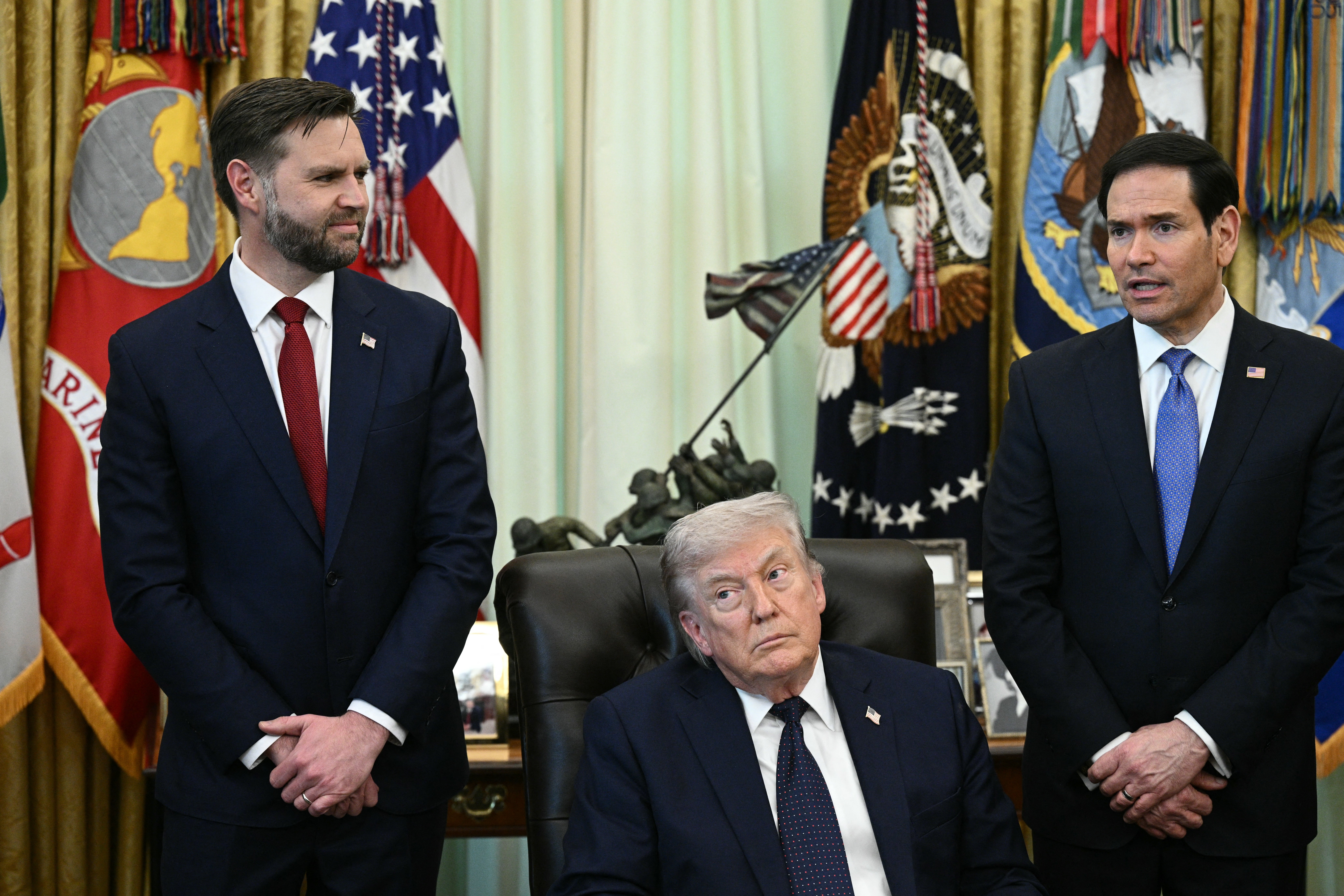 Tre menn i dress står i et offisielt rom med militærflagg