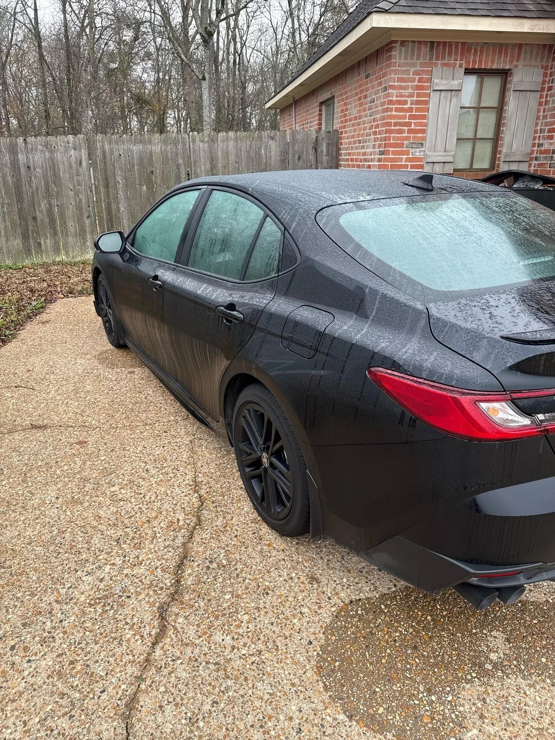 Sedan parked connected  a gravel driveway adjacent   a ceramic  location   with woody  fence. Overcast time  with disposable   histrion   branches successful  the background