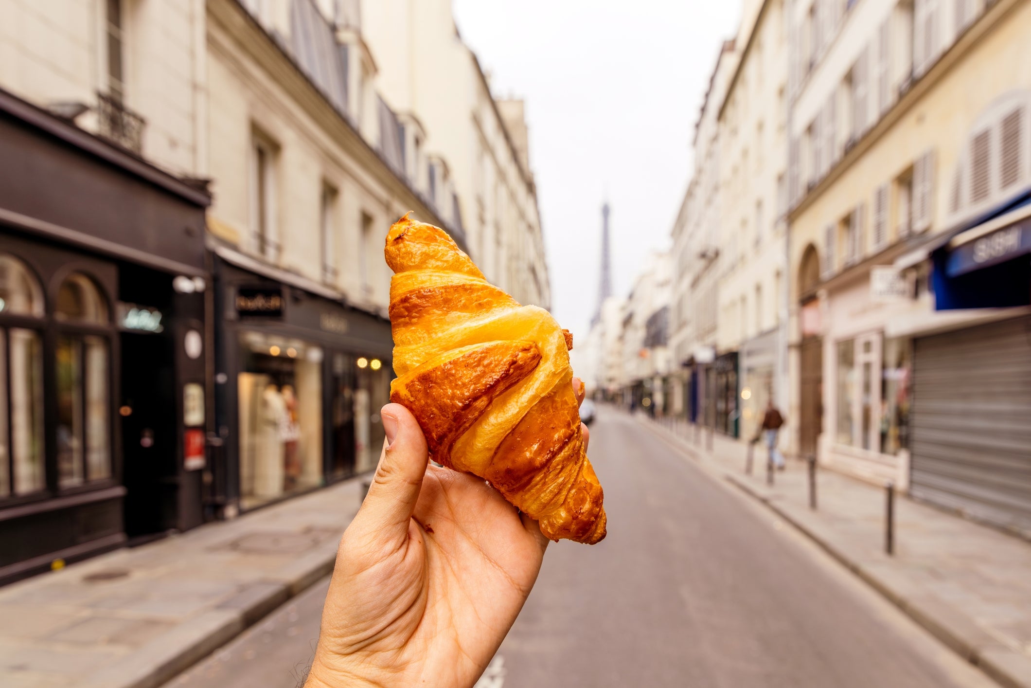 Hand holding a croissant on a quiet Paris street, with shops and the Eiffel Tower in the background
