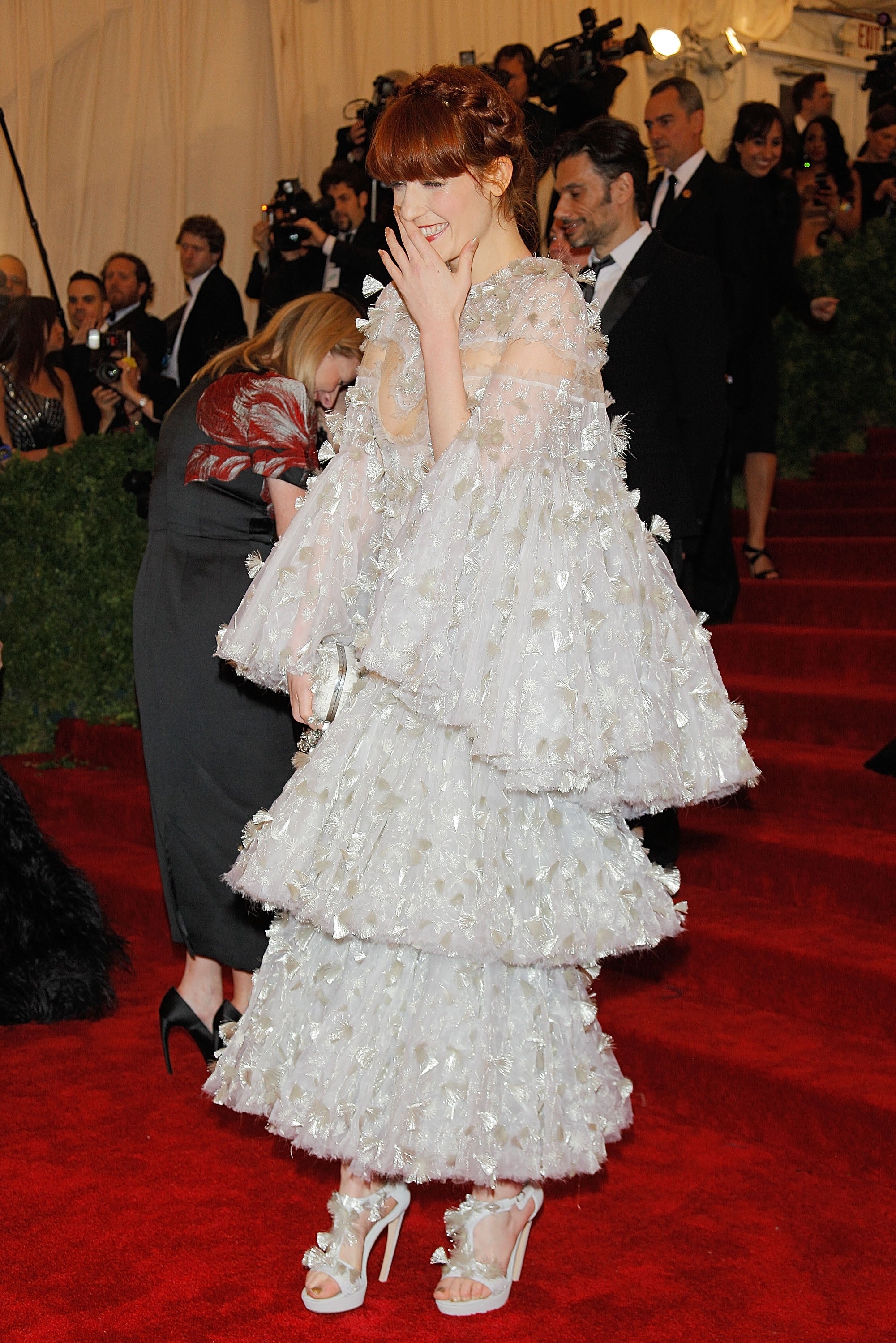 Person successful  layered, ruffled gown with floral details connected  a reddish  carpet, smiling with manus  adjacent   face, surrounded by photographers and attendees