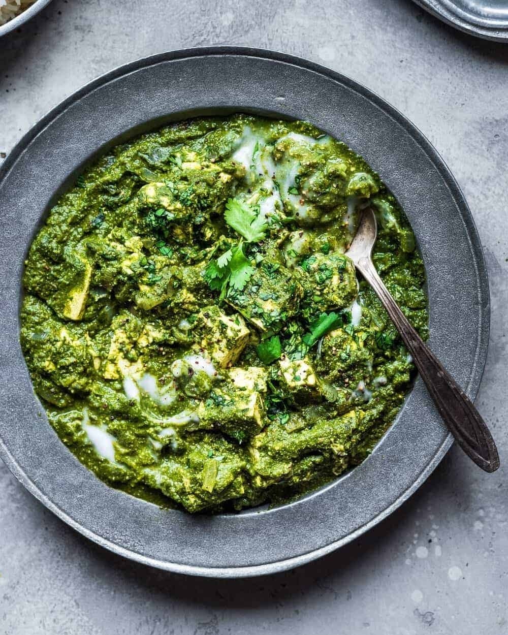 A bowl of palak paneer garnished with cilantro and a drizzle of cream, served in a gray dish with a spoon