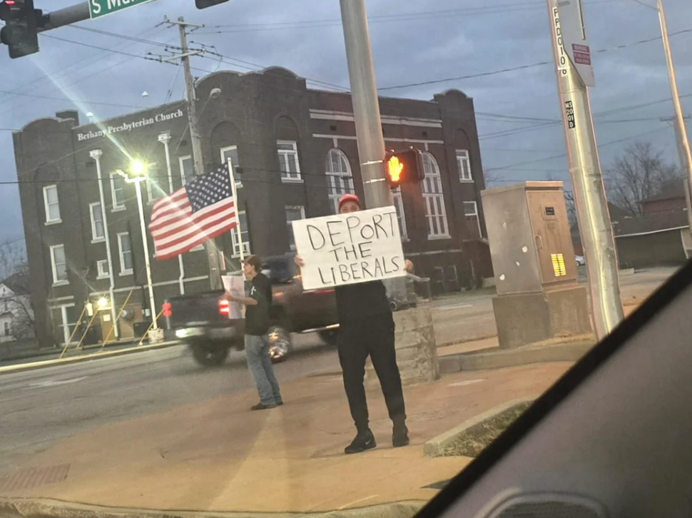 A idiosyncratic   stands connected  a thoroughfare  holding a motion   that reads "Deport the Liberals" adjacent   a religion  and a idiosyncratic   with an American flag