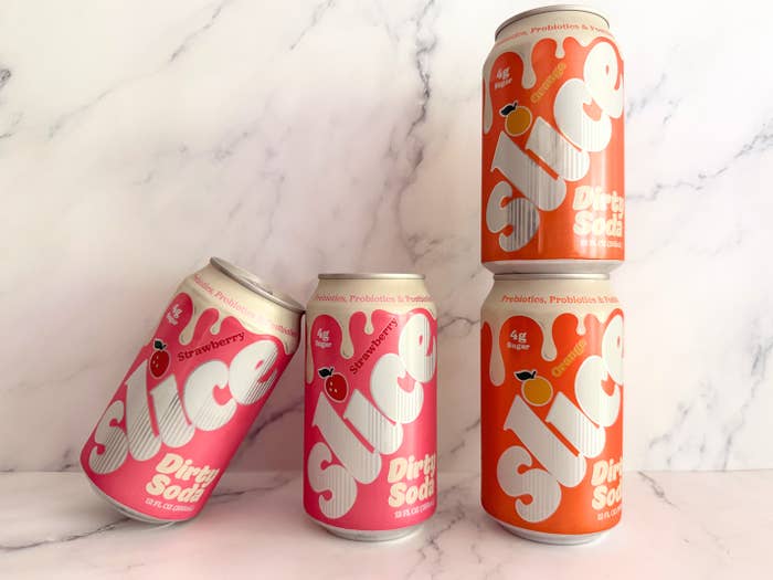 Four cans of Slice dirty soda, two strawberry-flavored and two orange-flavored, are arranged on a marble surface