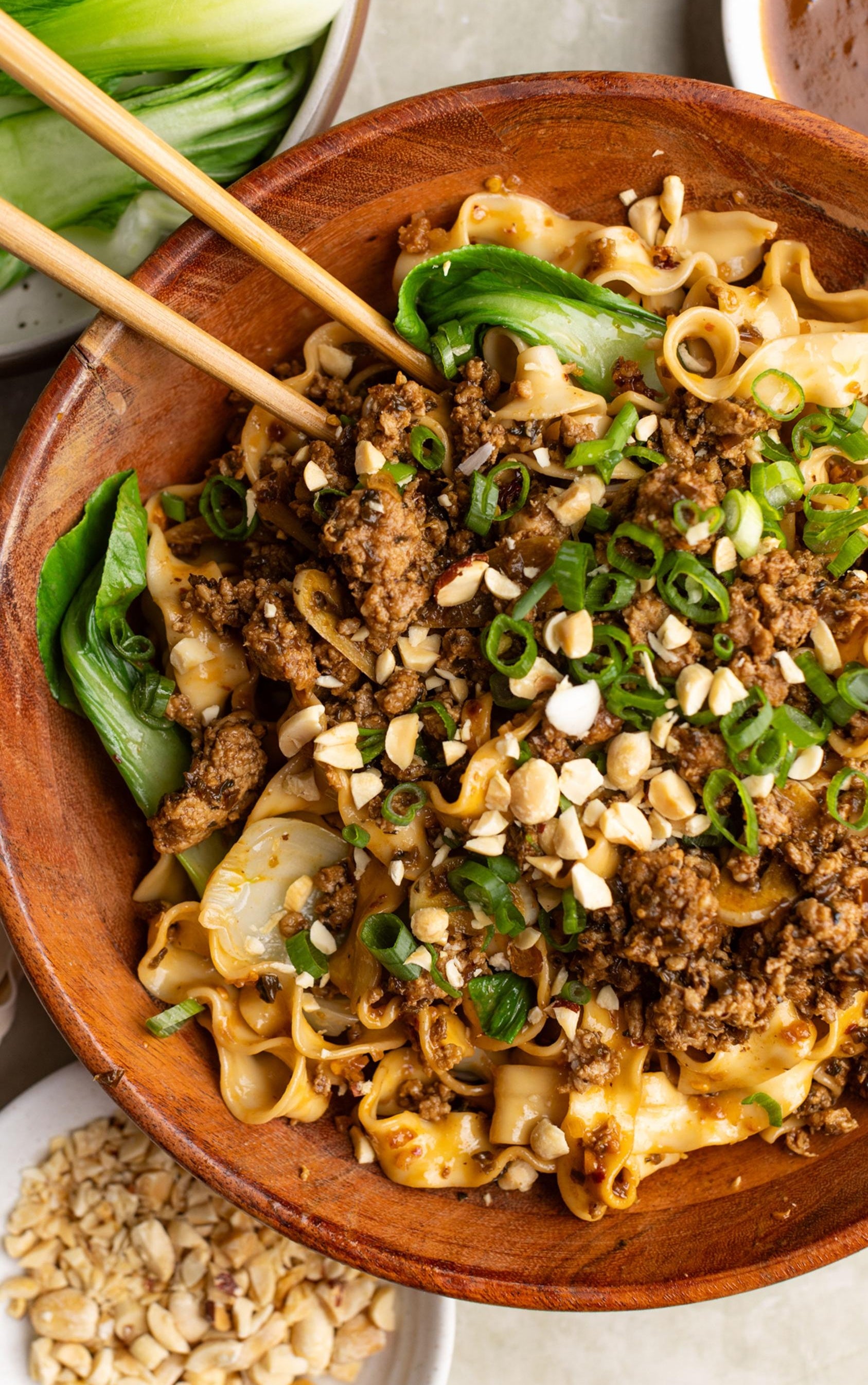 Bowl of noodles topped with minced meat, chopped peanuts, and green onions, with chopsticks and bok choy visible
