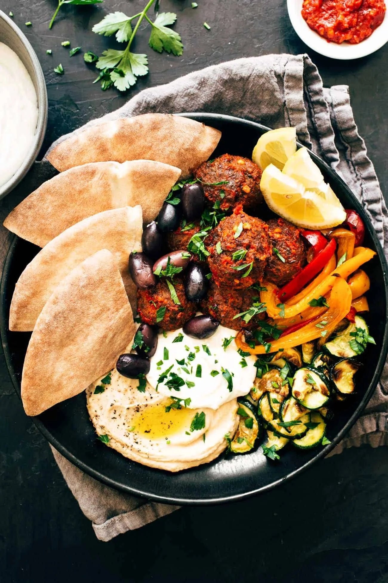A plate with hummus, pita, falafel, olives, grilled vegetables, and lemon wedges, garnished with herbs