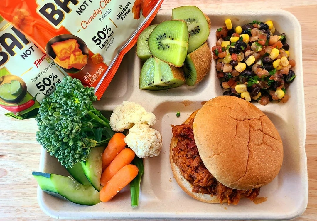 Tray with a pulled pork sandwich, broccoli, carrots, cauliflower, cucumber, sliced kiwi, maize  salsa, and chips successful  a schoolhouse  cafeteria