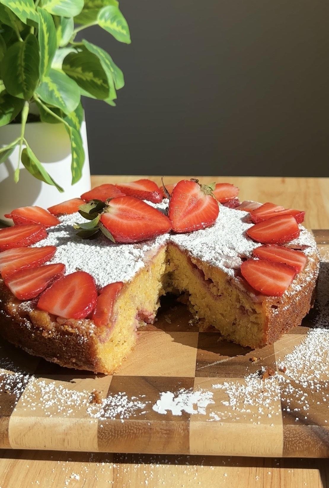 Strawberry-topped cake with a slice missing, dusted with powdered sugar on a wooden board next to a potted plant