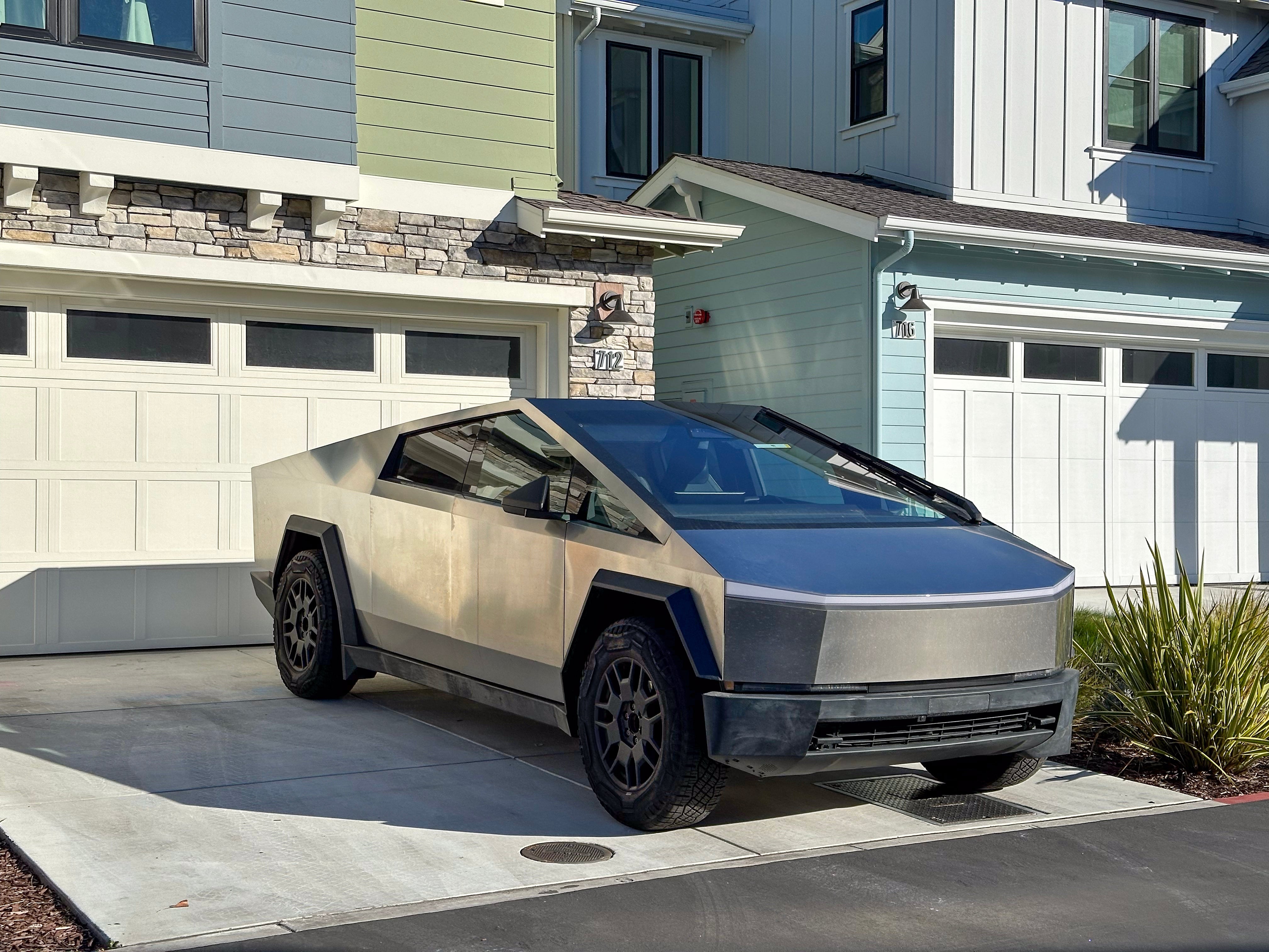 Futuristic, angular conveyance  resembling a Tesla Cybertruck parked successful  a residential driveway with modern   houses successful  the background