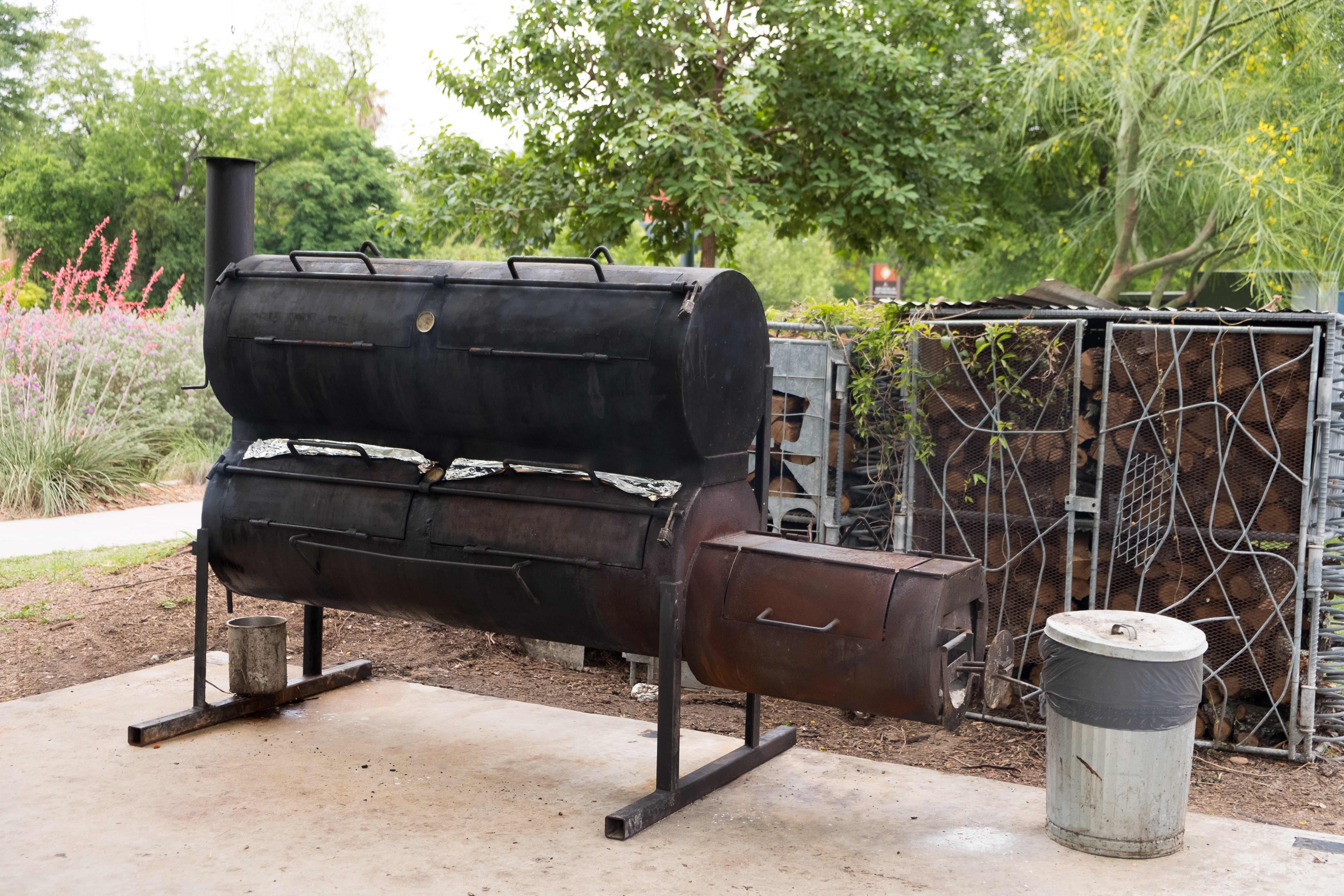 Outdoor metallic  barbecue smoker with a chimney and adjacent    woodpile amidst greenery, suggesting mentation  for grilling