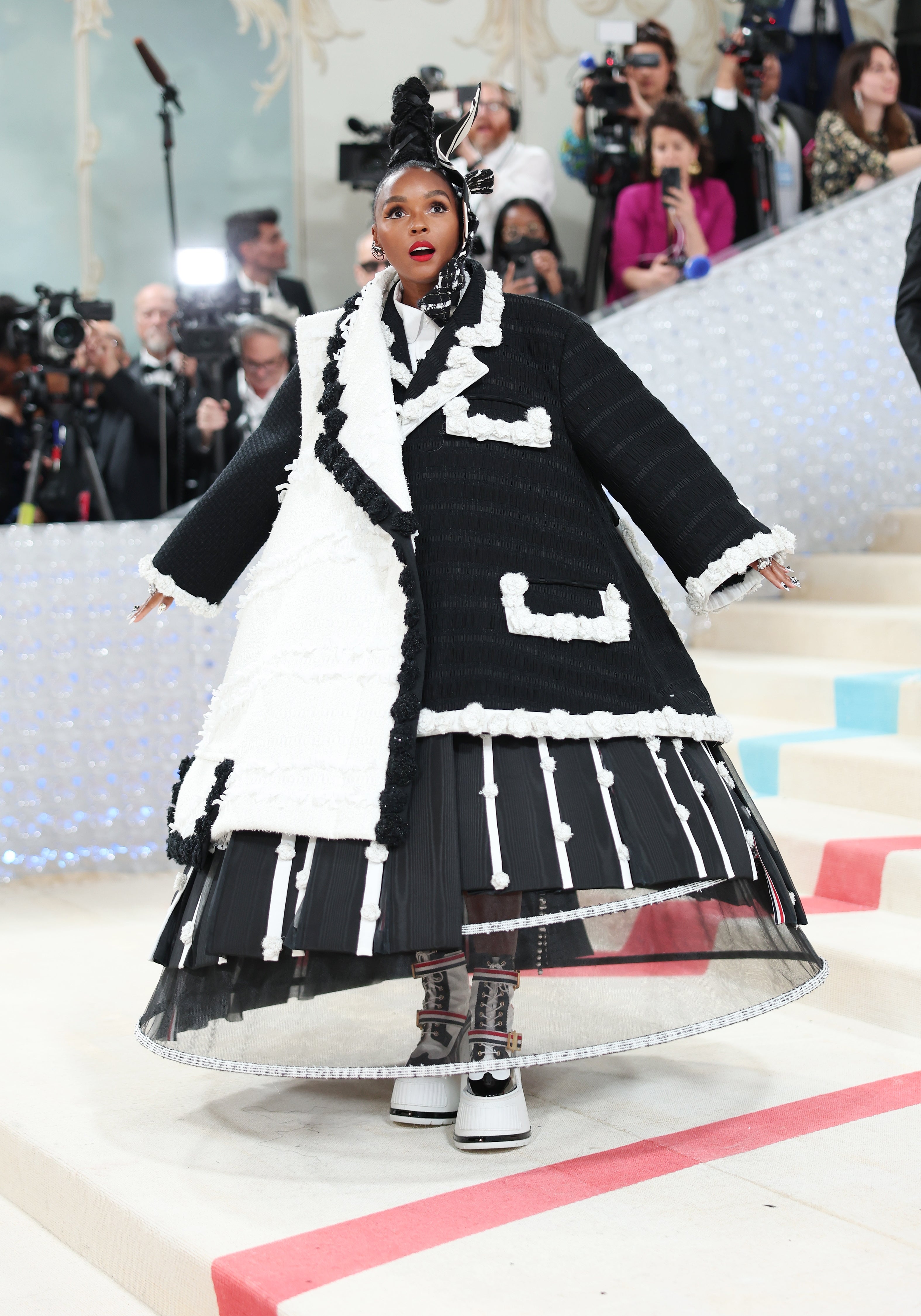 Person wearing a voluminous, avant-garde achromatic  and achromatic  overgarment  with intricate detailing connected  a reddish  carpet, surrounded by photographers