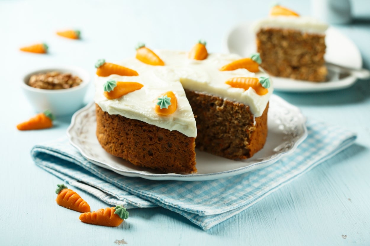 Carrot cake with cream cheese frosting, topped with decorative fondant carrots, displayed on a plate. One slice is cut and served nearby