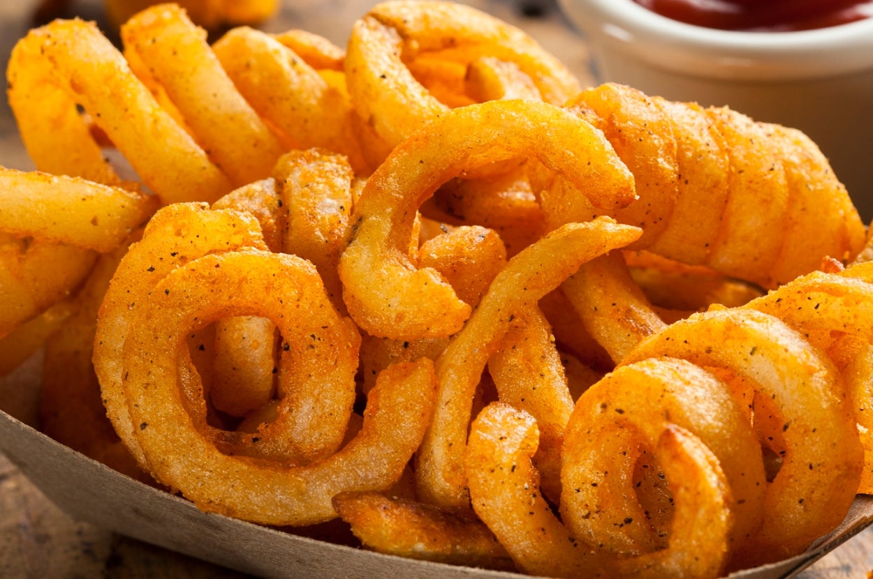 A close-up of seasoned curly fries in a tray, with a small bowl of sauce in the background