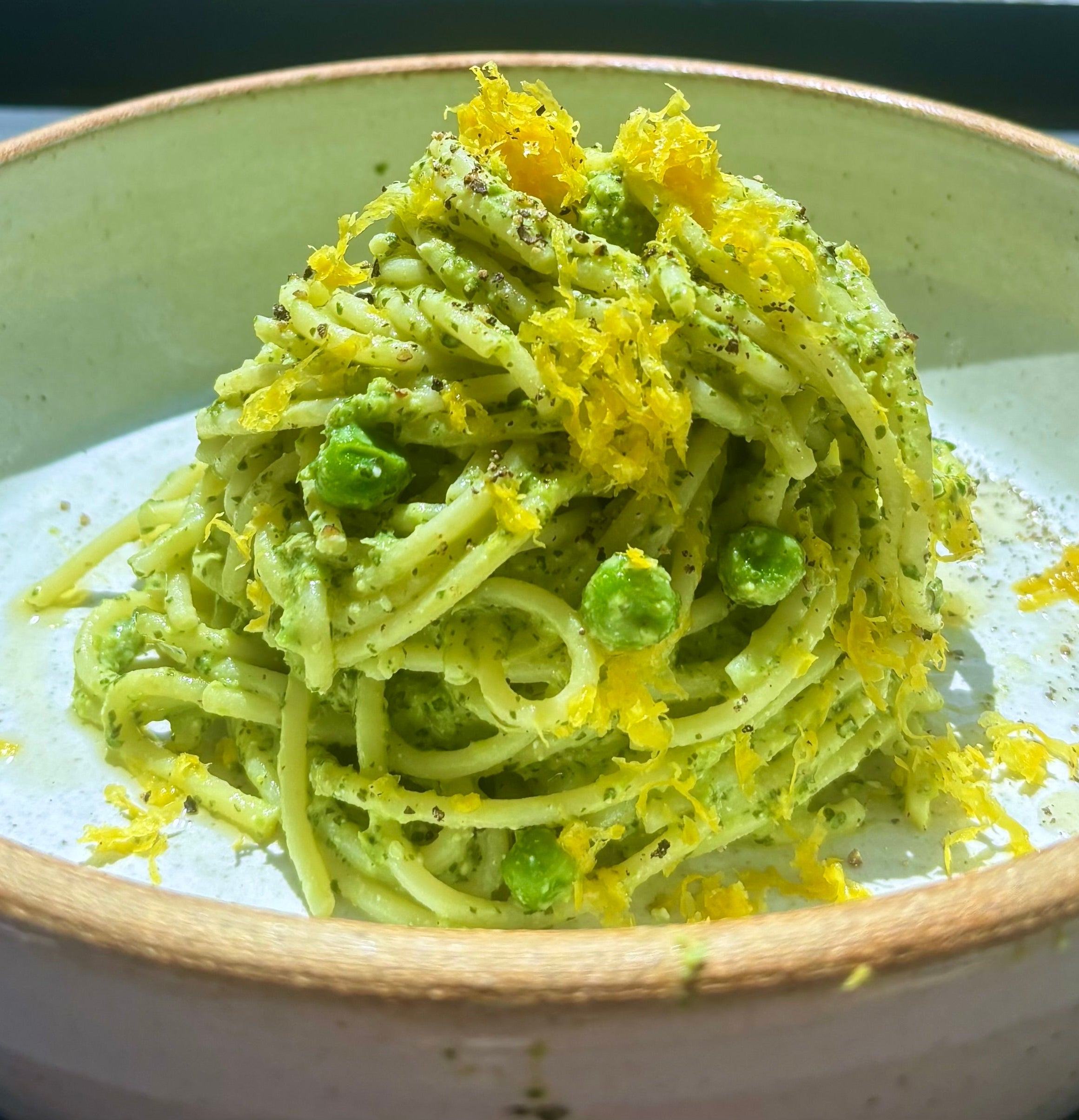 A bowl of spaghetti coated in green pesto sauce, garnished with peas and grated yellow zest, presented in a stoneware dish