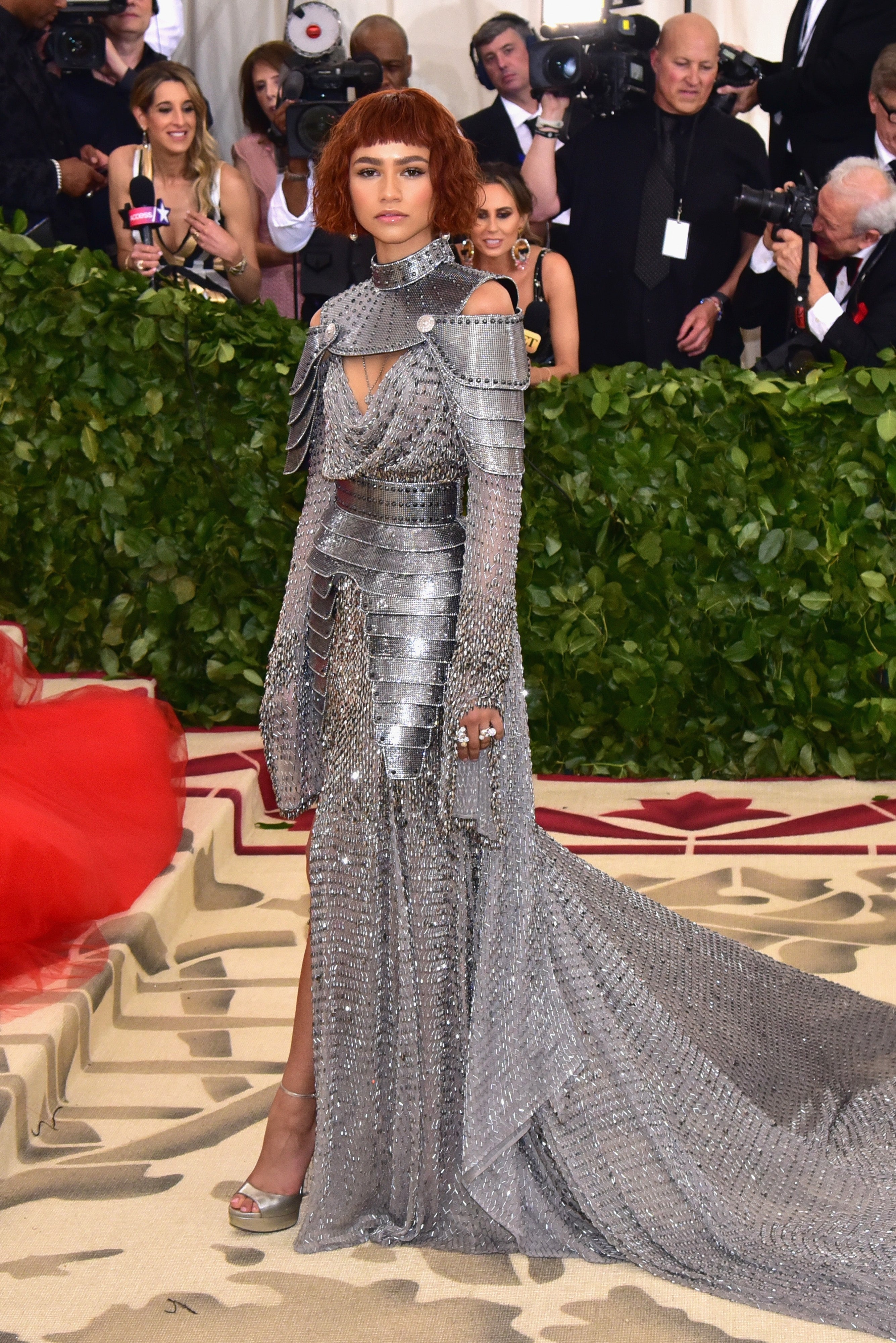 Person successful  metallic, armor-inspired gown with structured plan  connected  a ceremonial  lawsuit   reddish  carpet, surrounded by photographers and greenery backdrop