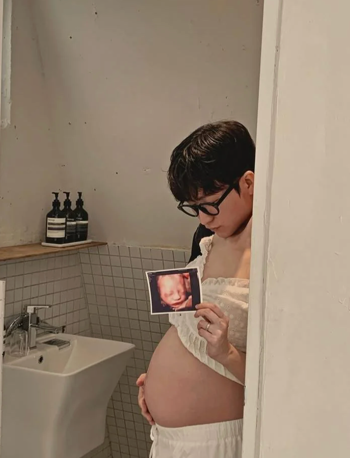 Pregnant person holding an ultrasound photo, standing in a tiled bathroom, looking at the image in their hand