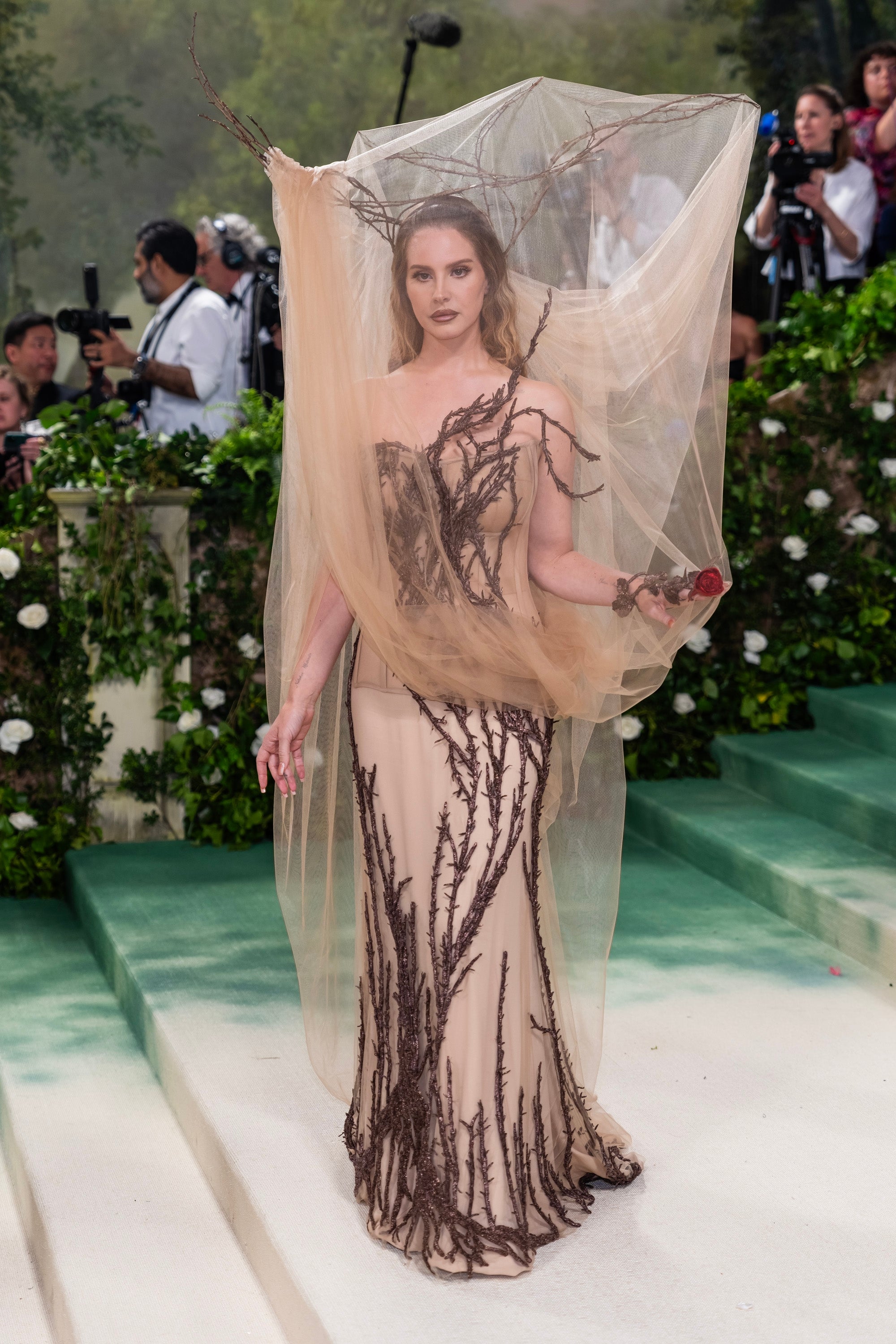 Person successful  an intricate gown with branch-like details and sheer veil connected  a greenish  staircase astatine  a ceremonial  event, surrounded by photographers