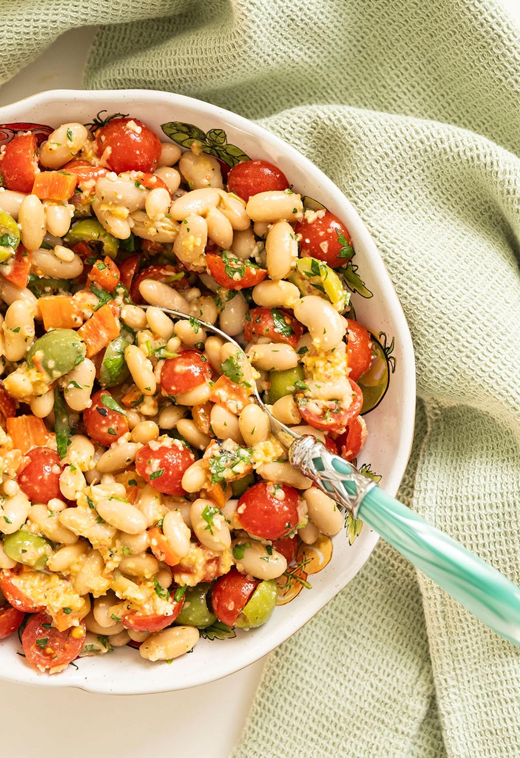 Bowl of mixed bean salad with tomatoes, avocado, and herbs, served with a spoon, on a light textured cloth