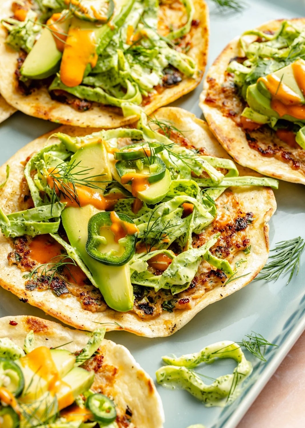 Flatbreads topped with avocado slices, shredded greens, and creamy dressing, garnished with dill on a light blue platter