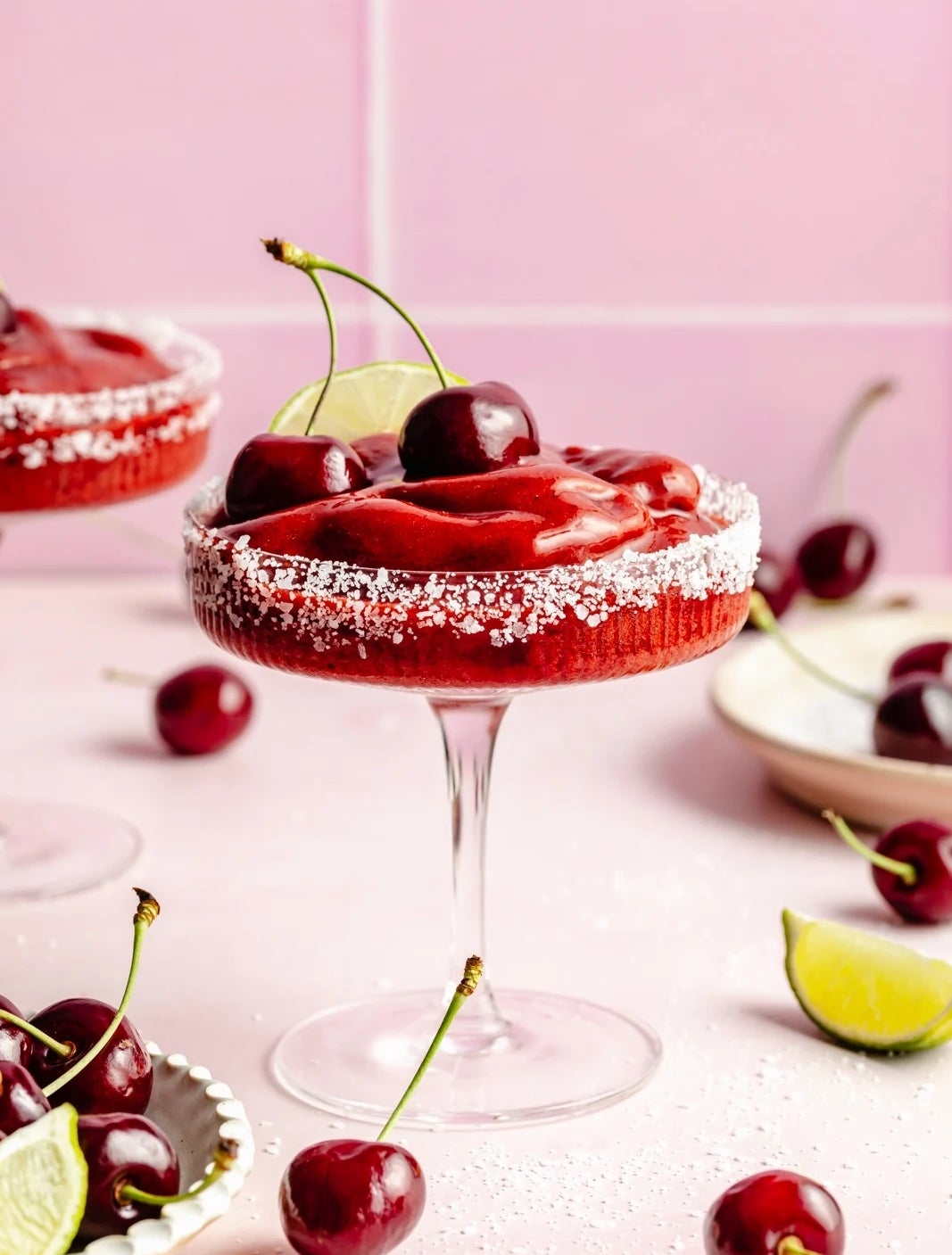 A frozen cherry cocktail garnished with fresh cherries and a lime slice, in a salt-rimmed glass, with more cherries and lime wedges nearby