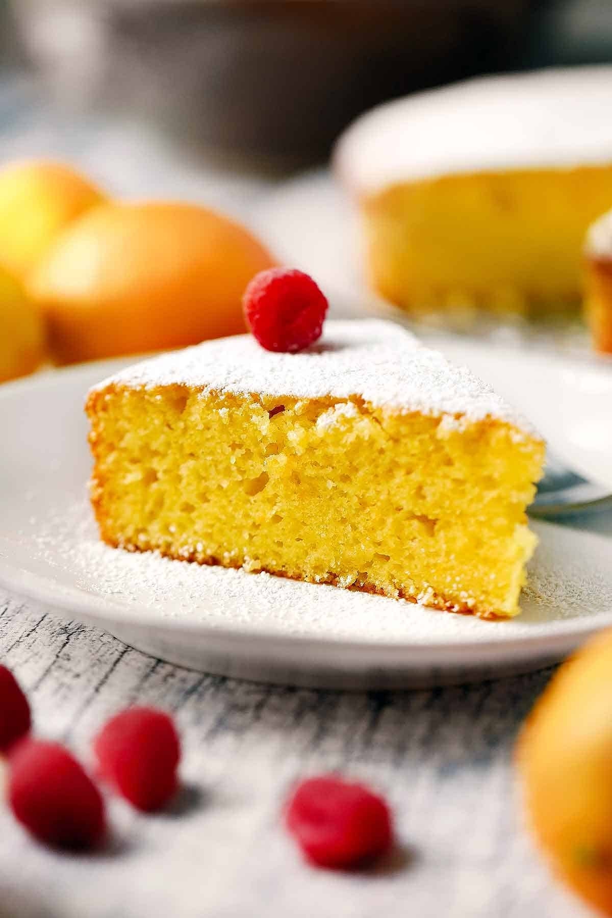 Slice of cake with powdered sugar and a raspberry on top, with more fruit in the background