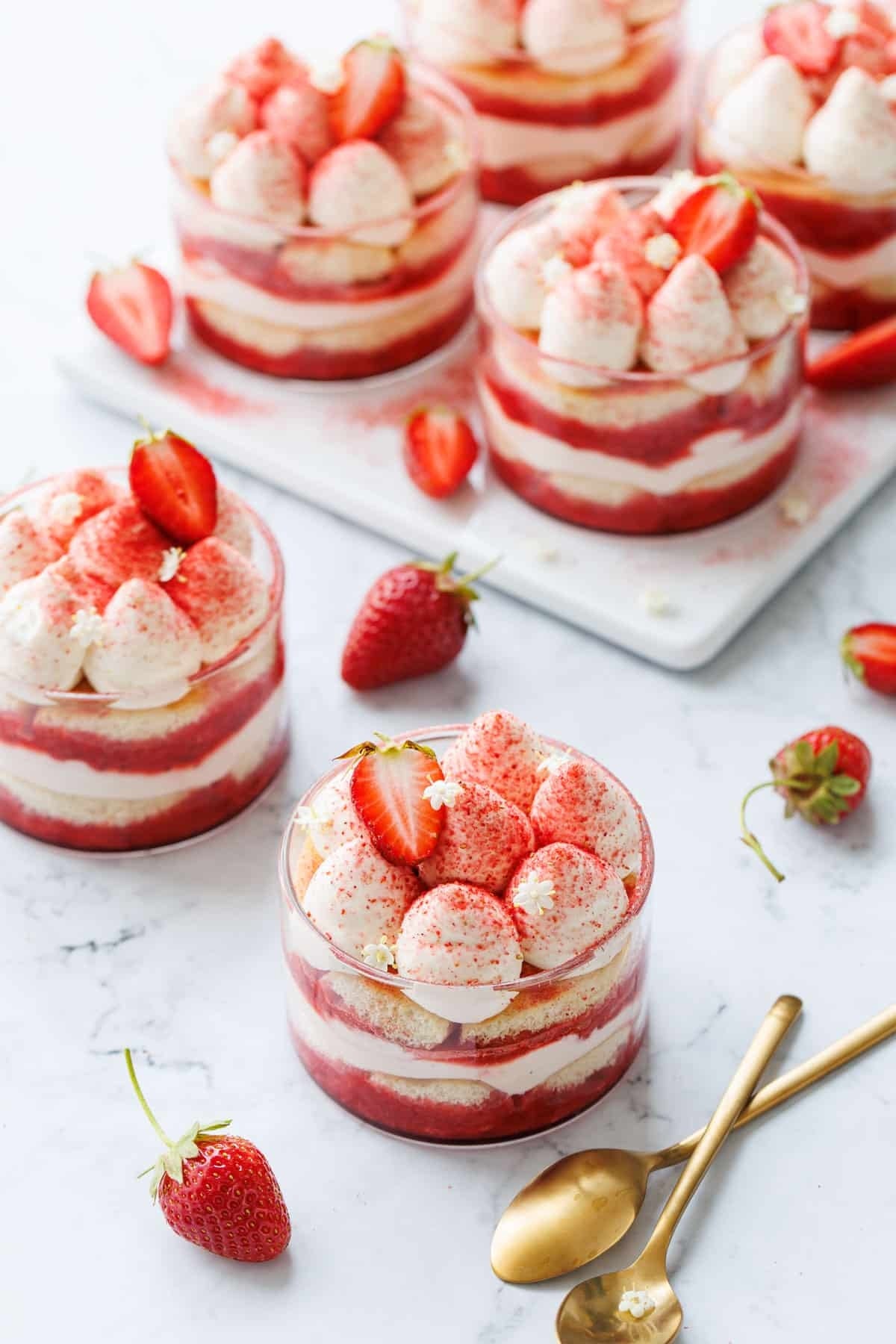 Glass cups filled with layers of whipped cream, strawberry slices, and sponge cake. Topped with whole strawberries and a dusting of powder