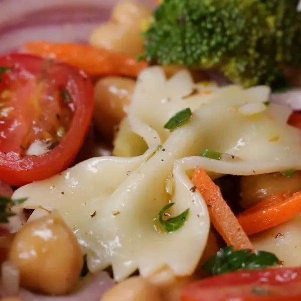 A close-up of a fresh pasta salad with bowtie pasta, cherry tomatoes, chickpeas, carrots, broccoli, and herbs