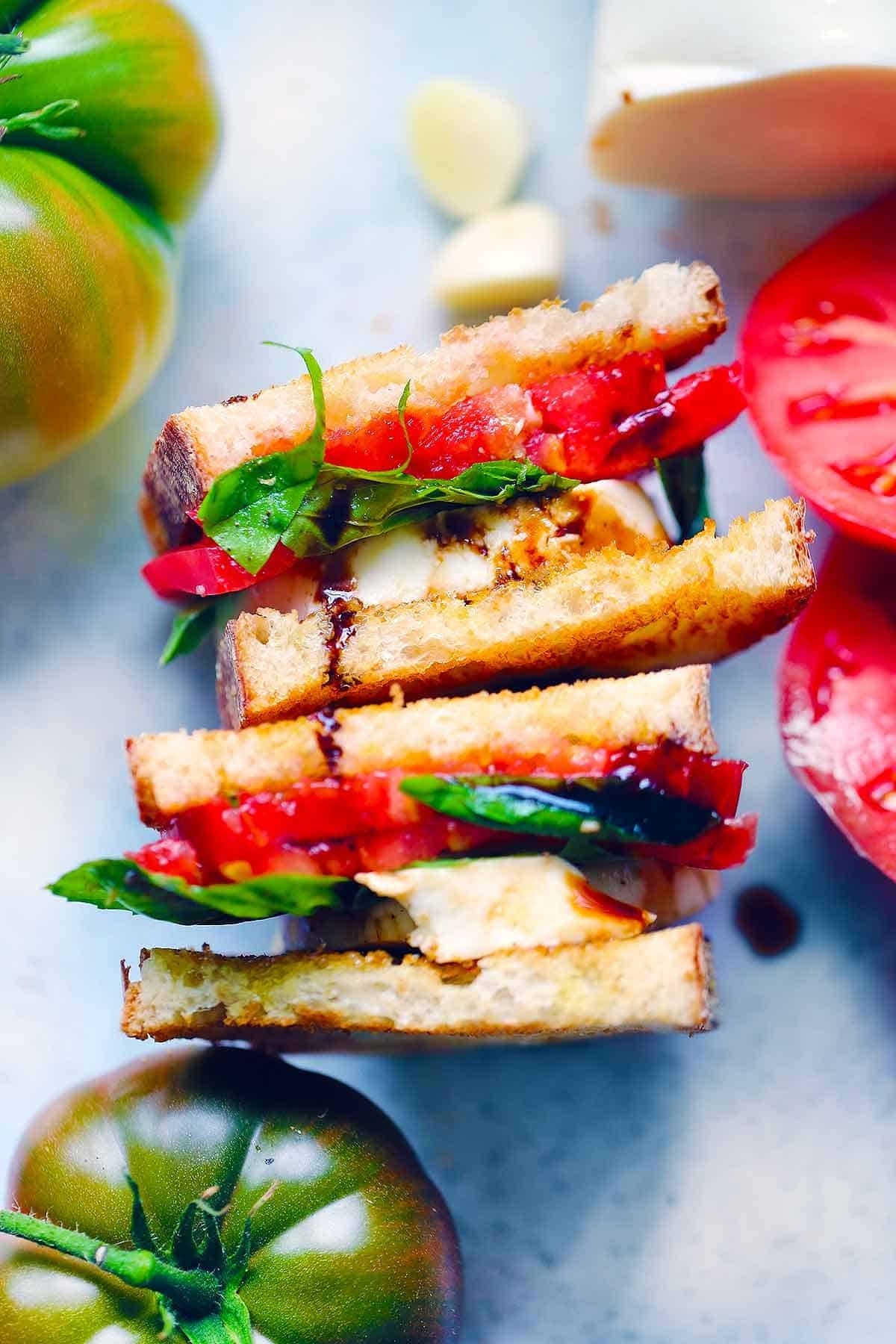 A stacked Caprese sandwich with mozzarella, tomato, basil, and balsamic glaze on toasted bread, surrounded by fresh tomatoes