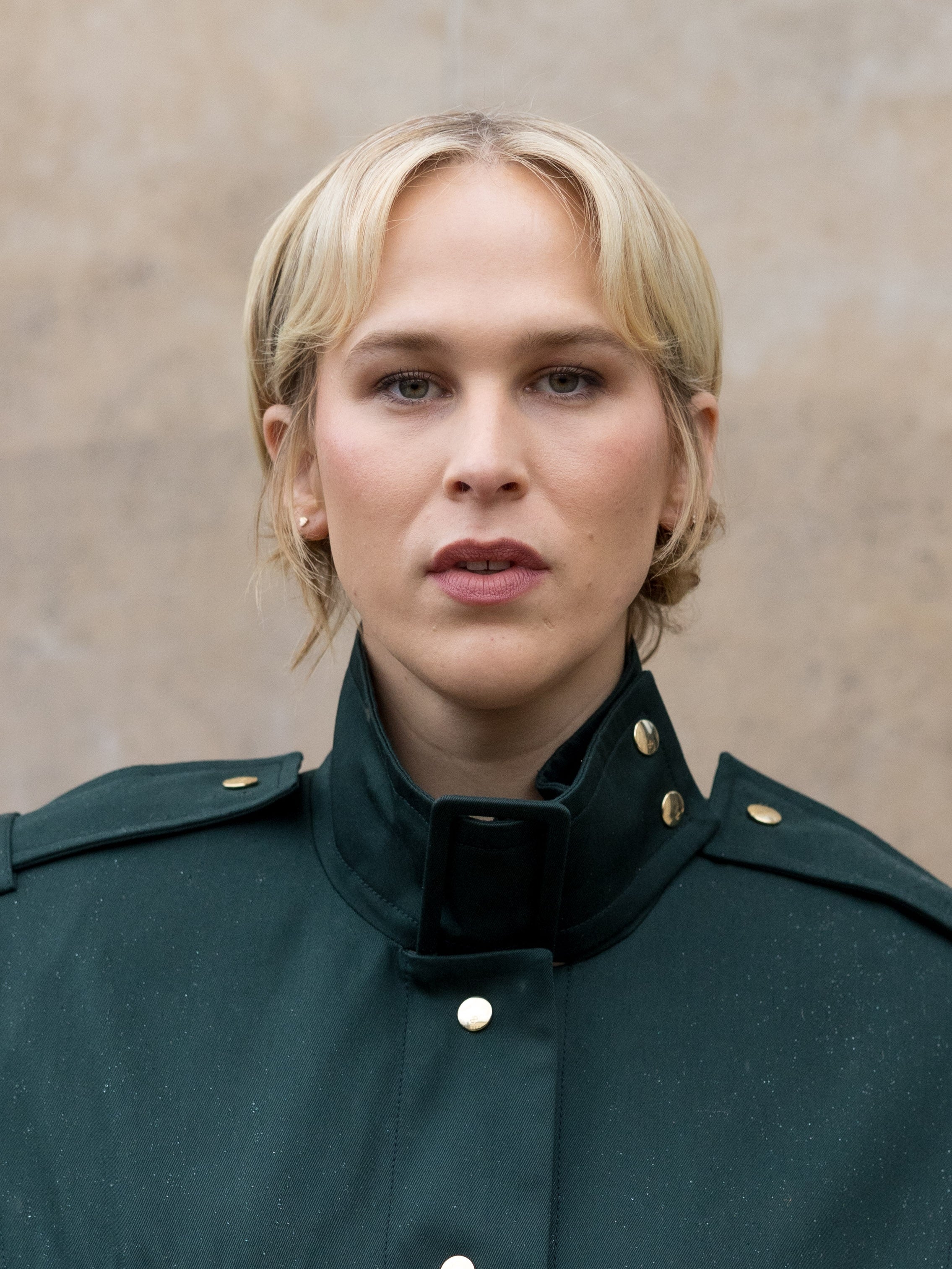Tommy Dorfman with short hair wearing a high-collared jacket with gold buttons, looking directly at the camera