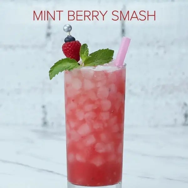Tall glass with a pink drink, crushed ice, mint leaves, raspberry, and a pink straw. Text reads "MINT BERRY SMASH."