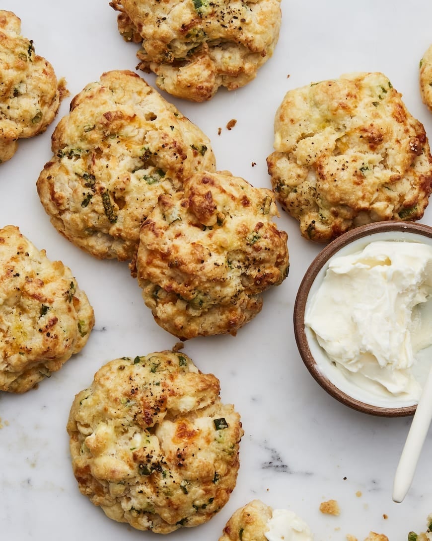 Biscuits with herbs and cheese on a marble surface, accompanied by a bowl of cream cheese with a spreader