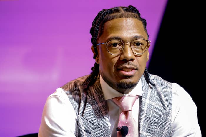 Nick Cannon successful  glasses with braided hair, wearing a plaid vest implicit    a garment  with a dotted tie, seated against a plain background