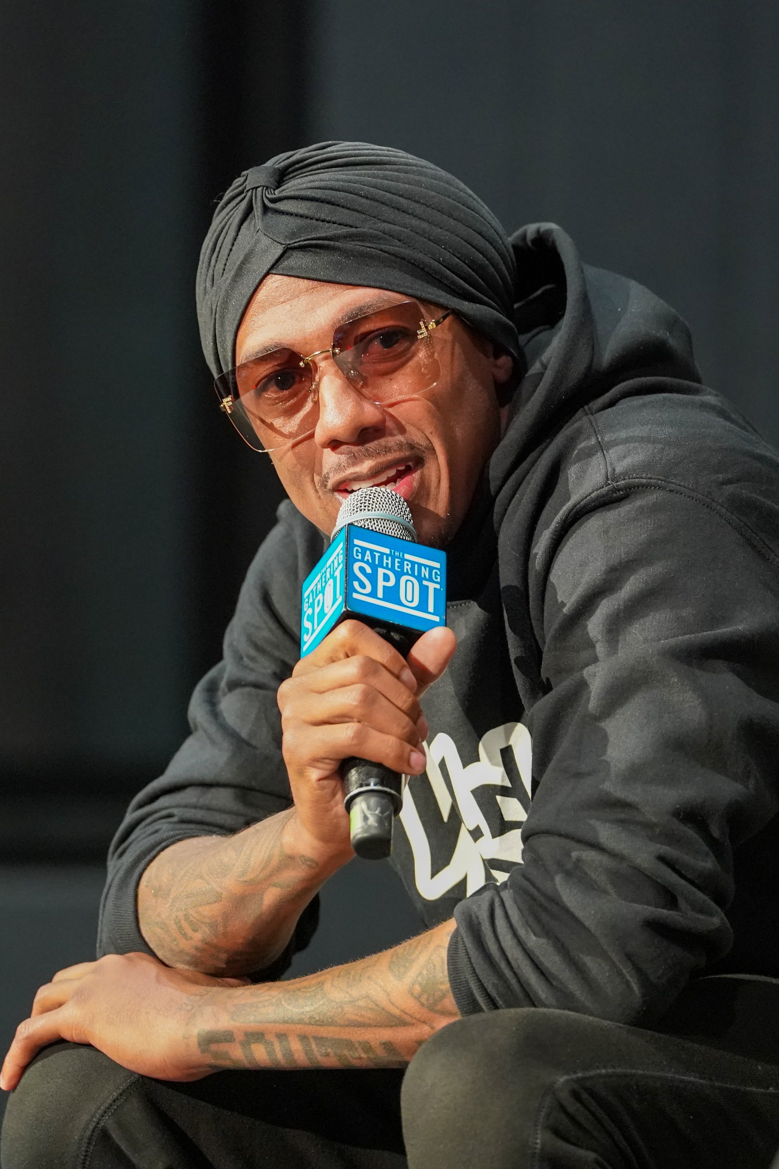 Nick Cannon wearing a hoodie and a turban speaks into a microphone astatine  an event, seated and wearing sunglasses