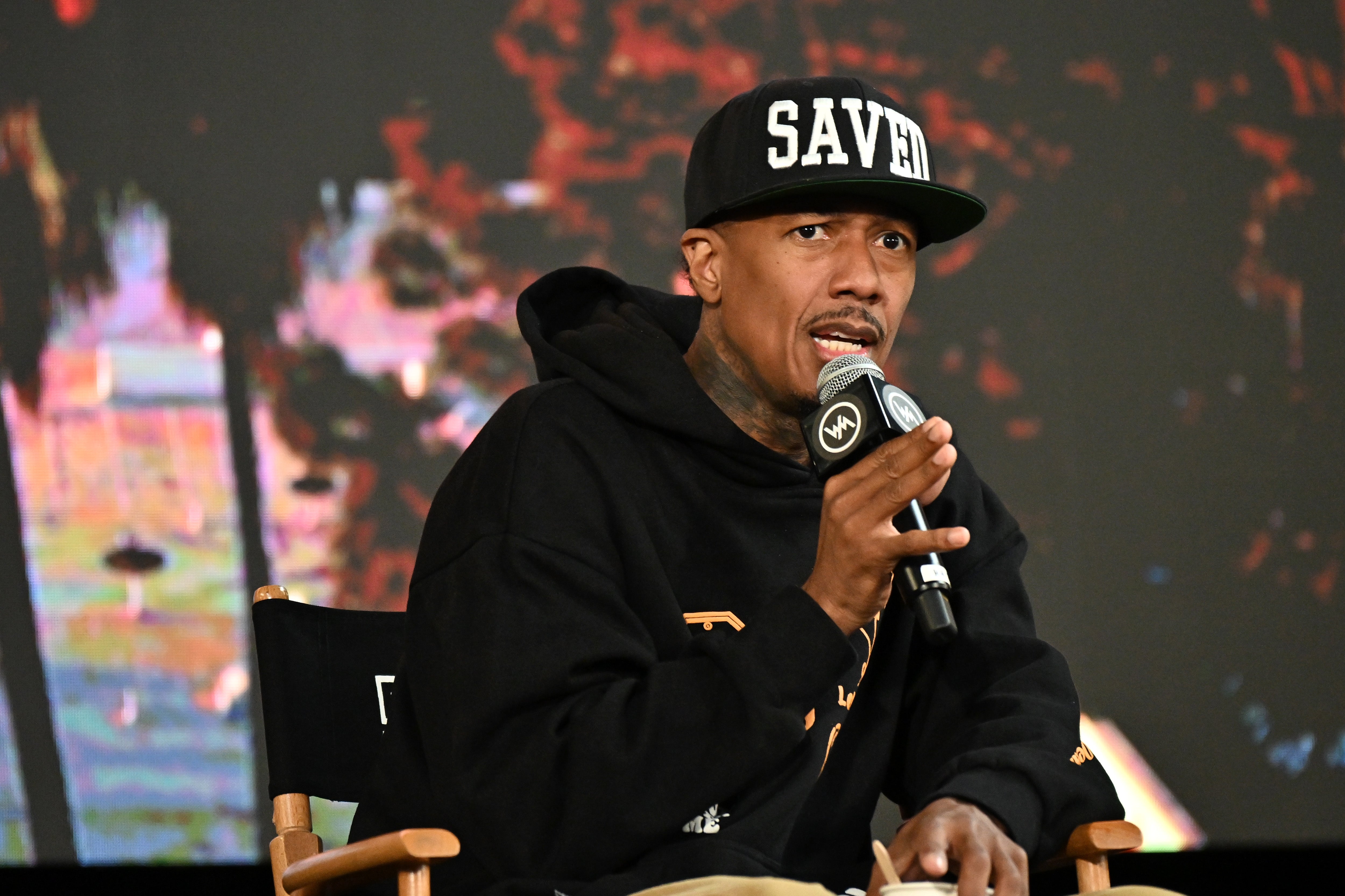 Nick Cannon successful  a "SAVED" headdress  and hoodie speaks into a microphone portion    seated successful  a director’s seat  astatine  an event