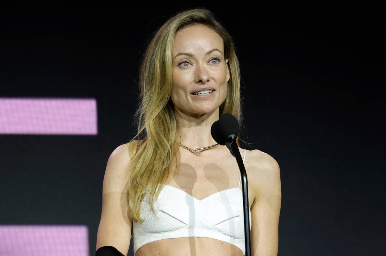 Olivia Wilde at a podium, wearing a stylish strapless top, is speaking into a microphone at a public event