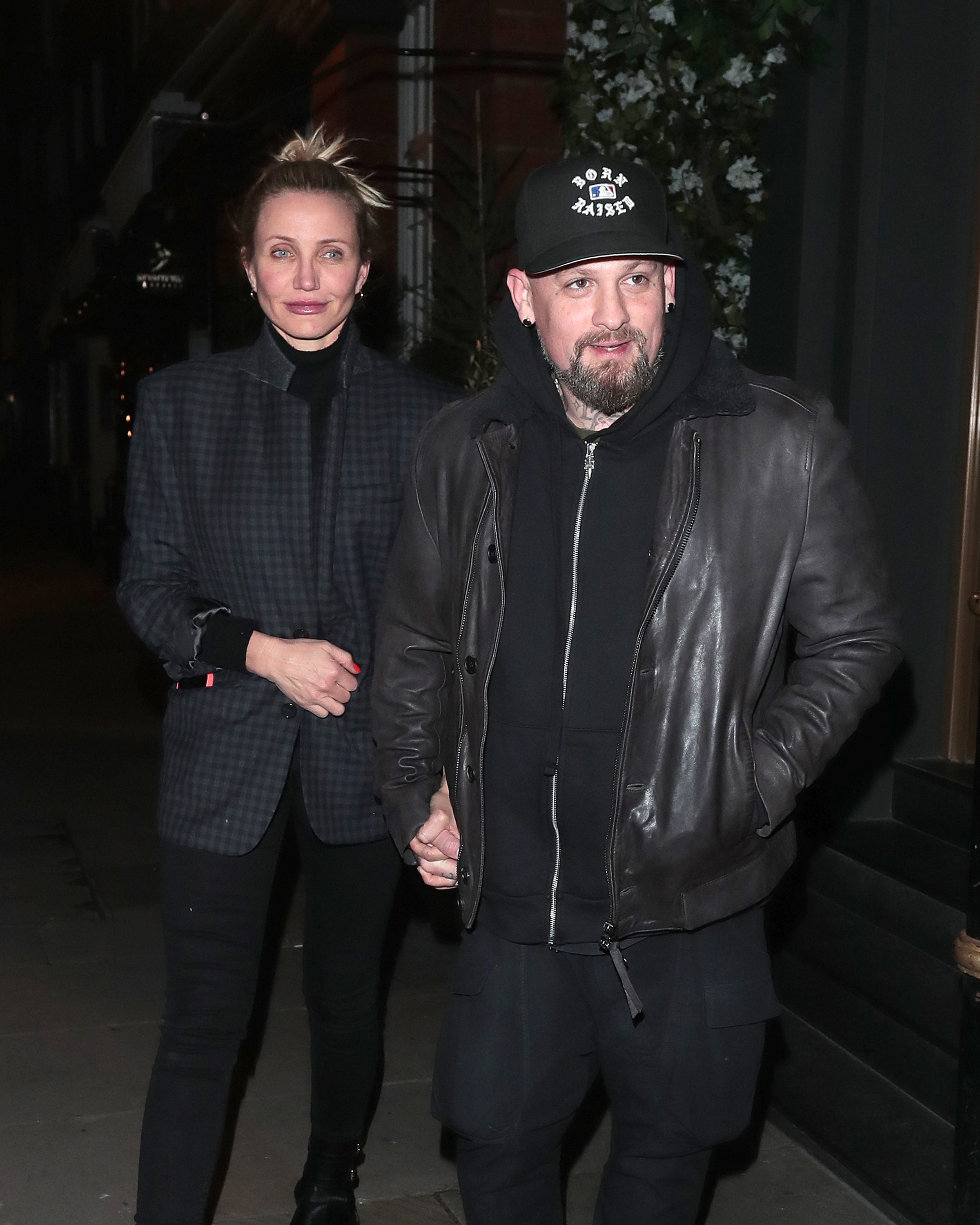 Two radical   walking hand-in-hand extracurricular  astatine  night; 1  wears a checked blazer, the different   a leather overgarment   and cap