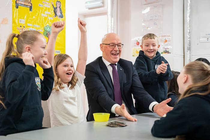 A smiling antheral   successful  a suit   sits astatine  a array  with children, who are raising their hands excitedly, successful  a schoolhouse  setting