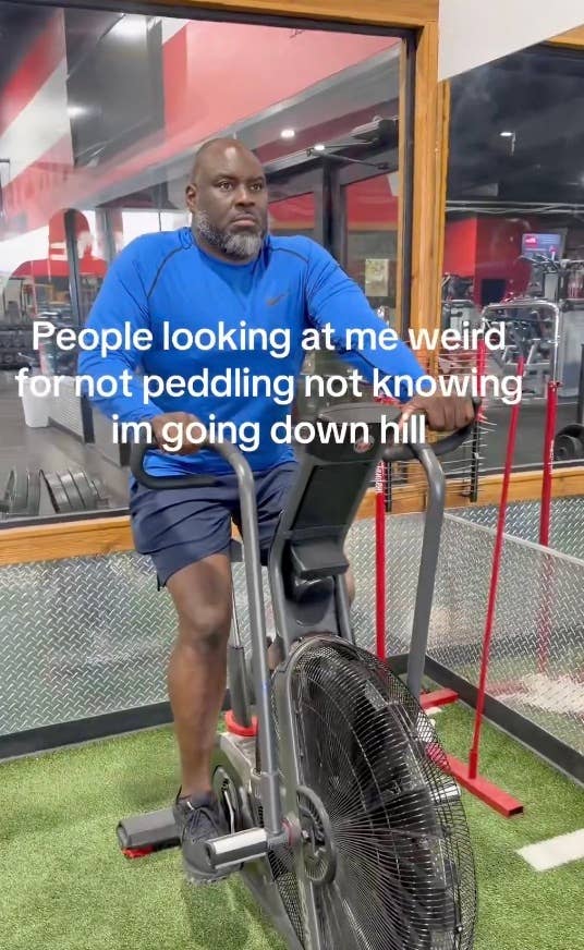 Man sitting on a stationary bike in a gym with text overlay reading, "People looking at me weird for not pedaling not knowing I'm going downhill."