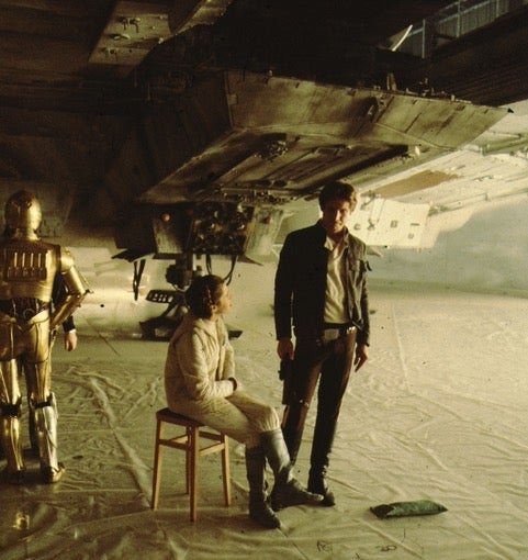 Actors connected  a movie  set, with 1  seated and 2  standing, engaged successful  conversation. A sci-fi spacecraft is partially disposable   successful  the background