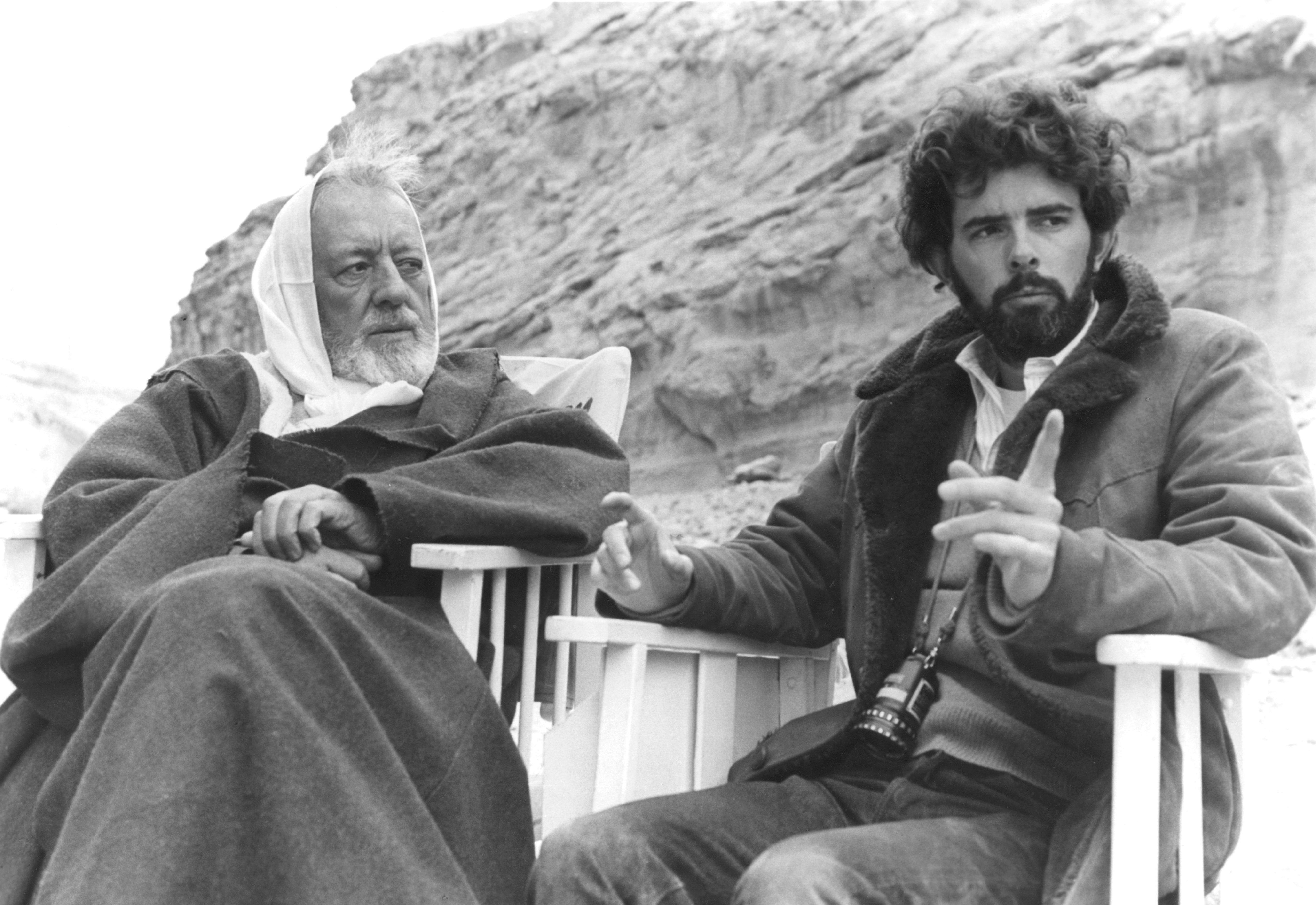 Two radical   beryllium   outdoors successful  beforehand   of a rocky backdrop. One gestures portion    talking, the different   listens attentively. One is successful  a robe, the different   successful  a coat