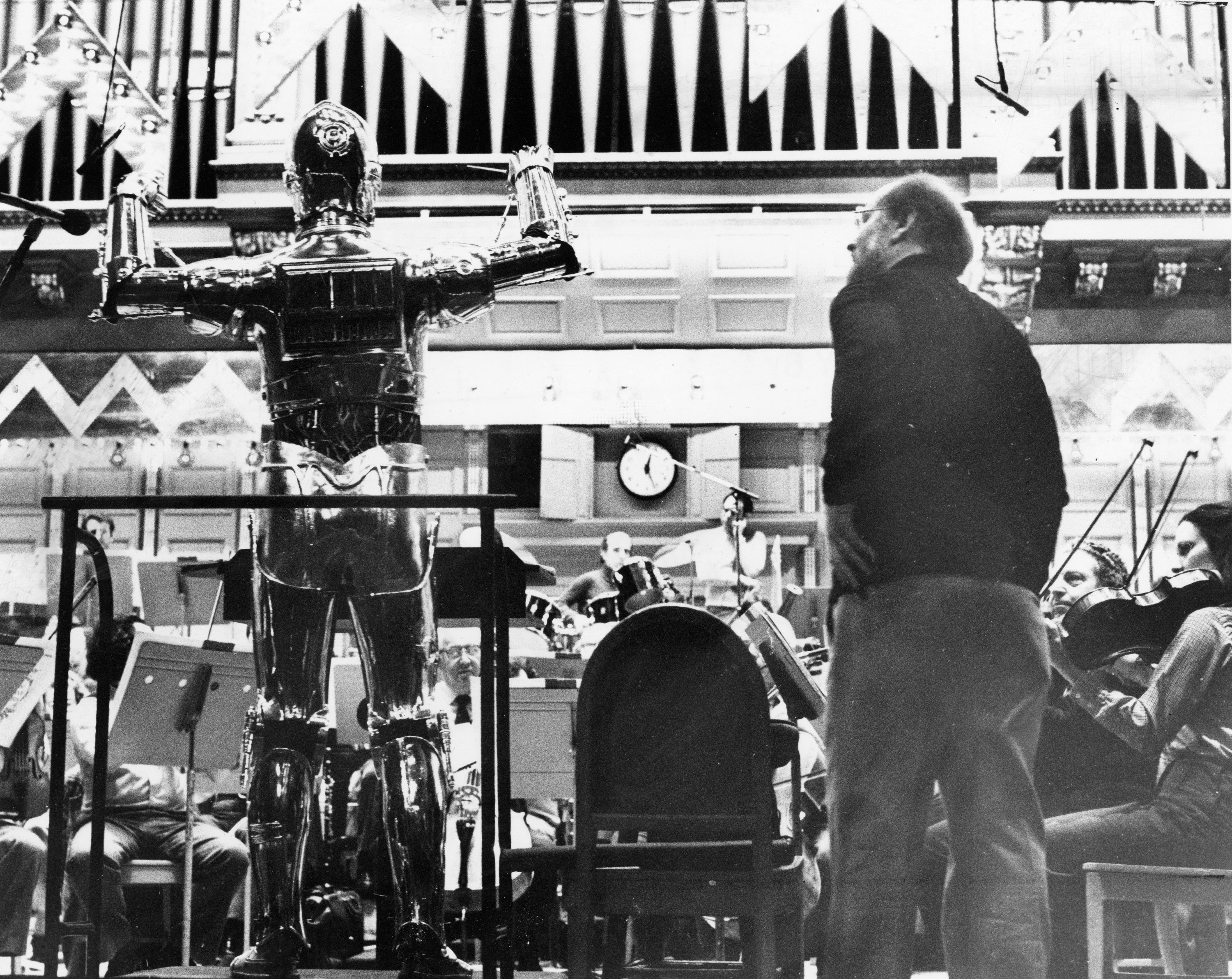 A conductor stands connected  a podium adjacent  to a idiosyncratic   successful  a robot costume, starring  an orchestra during a signaling  session