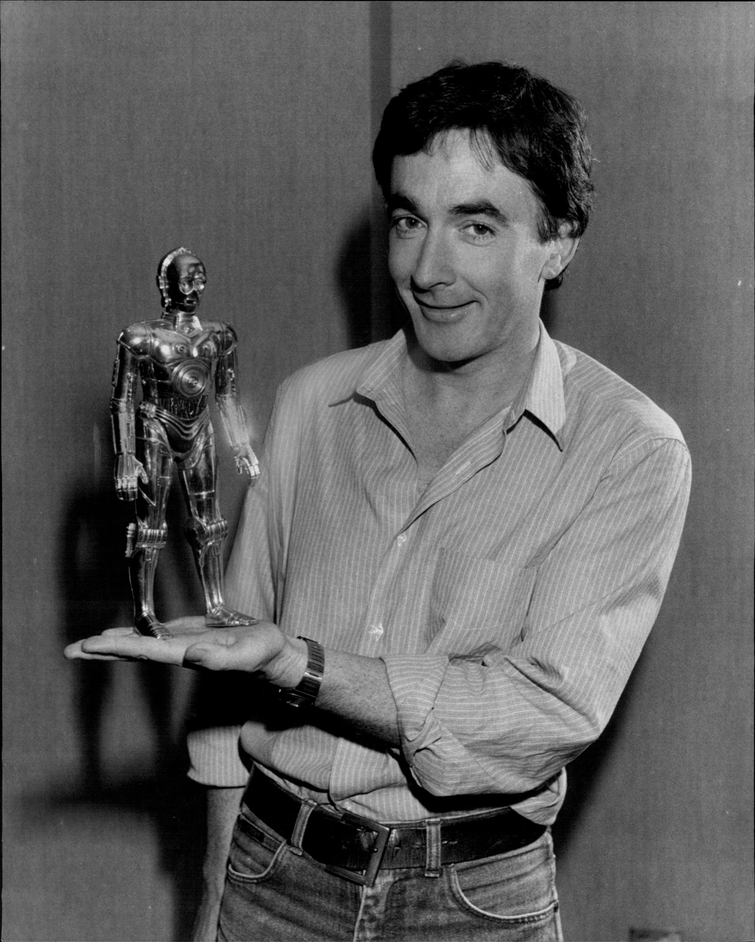 Person holding a tiny  metallic robot statue, smiling, wearing a casual garment  and jeans