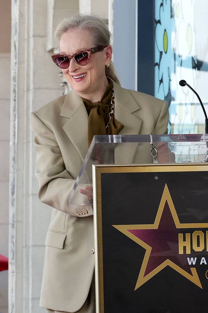 A idiosyncratic   speaks astatine  a podium during a Hollywood Walk of Fame event. They deterioration  an elegant beige suit   and chic sunglasses, with a joyful expression