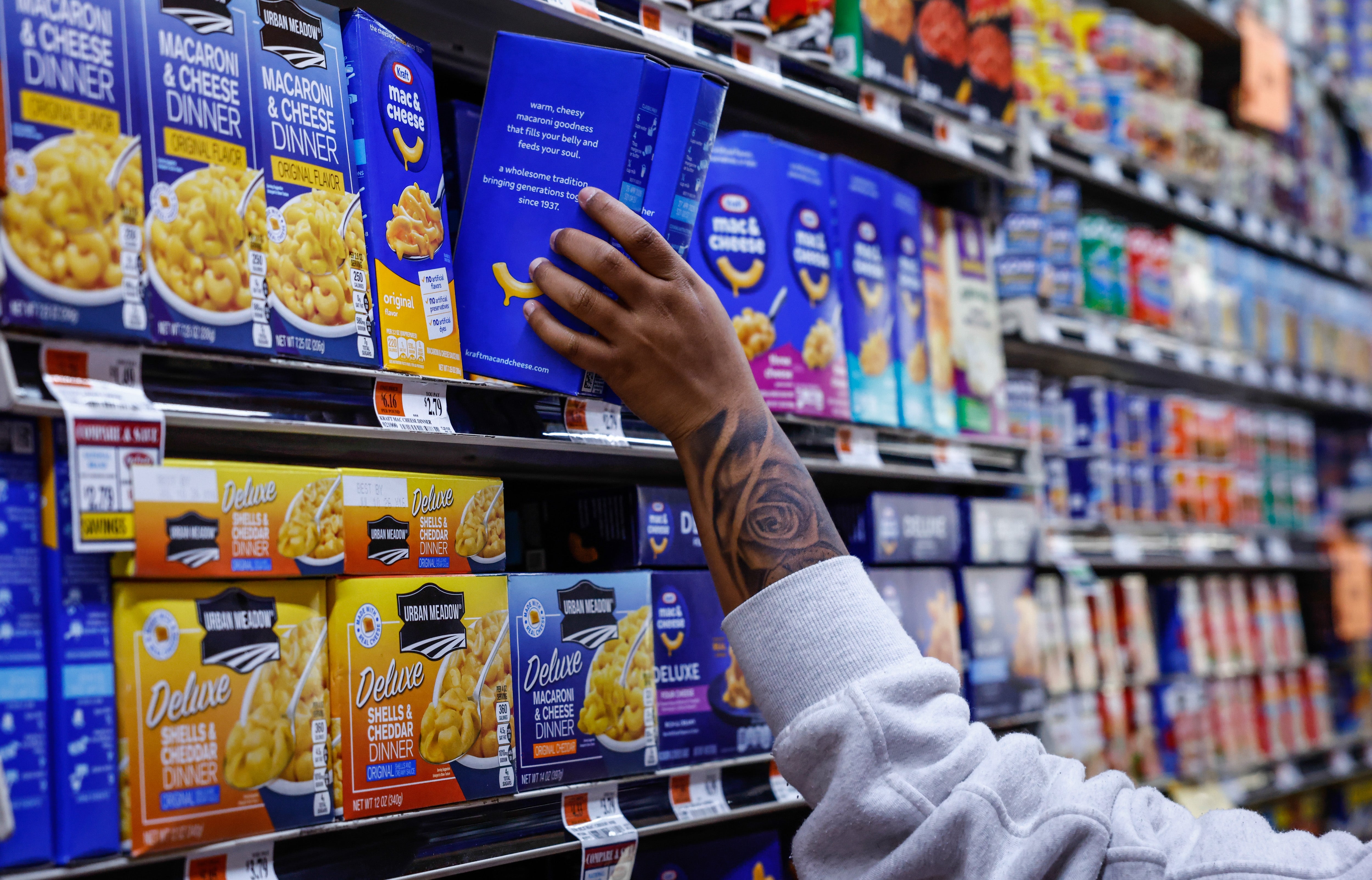 Person reaching for a container of macaroni and food connected a market store shelf. Multiple brands and types of pasta are displayed